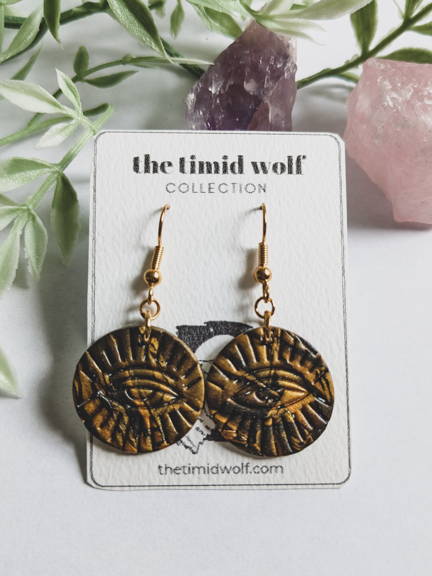 This photo shows a pair of black and gold polymer clay earrings in the shape of a circle. These earrings feature stainless steel hooks, and the design is of the "All-Seeing Eye" is stamped on the circle.